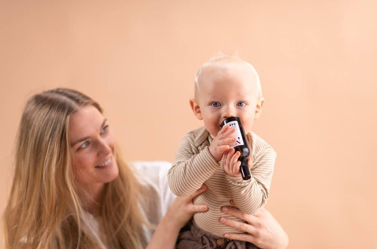 Smilende kvinde holder en baby, som har en flaske Økologisk Babyolie i hænderne, foran en ferskenfarvet væg