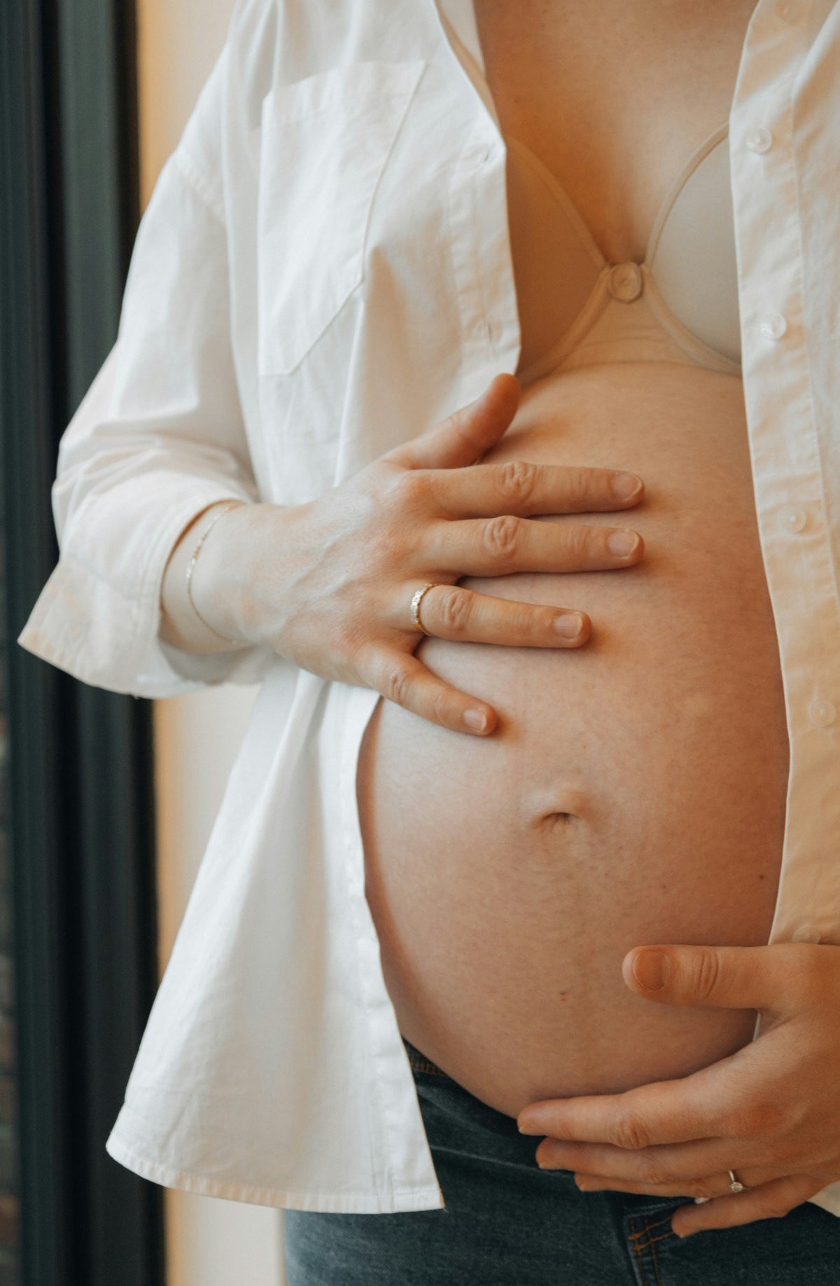Gravid kvinde i åben hvid skjorte med hænderne på maven i blødt vinduslys
