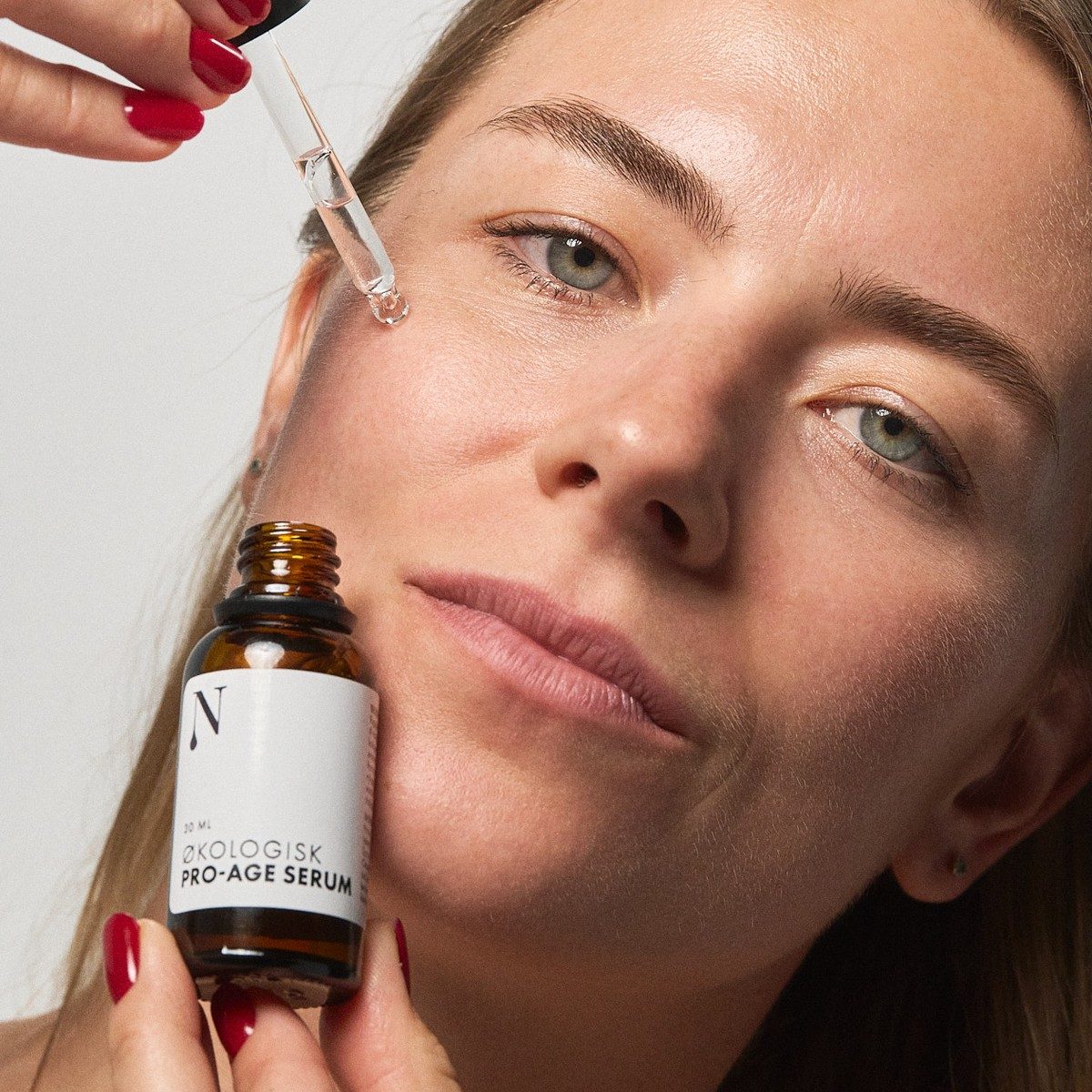 Kvinde drypper Pro-Age Serum på kinden med pipetten og holder flasken i den anden hånd